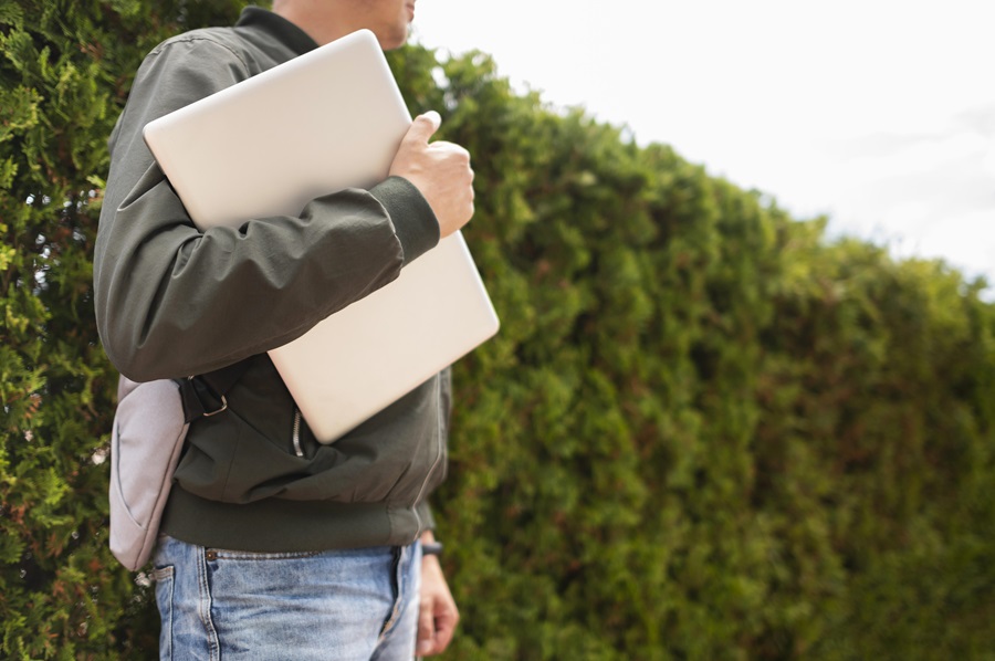 Um homem com postura confiante segura um tablet grande em meio a uma plantação do agronegócio; ele veste calça jeans, jaqueta esportiva e uma pequena mochila.