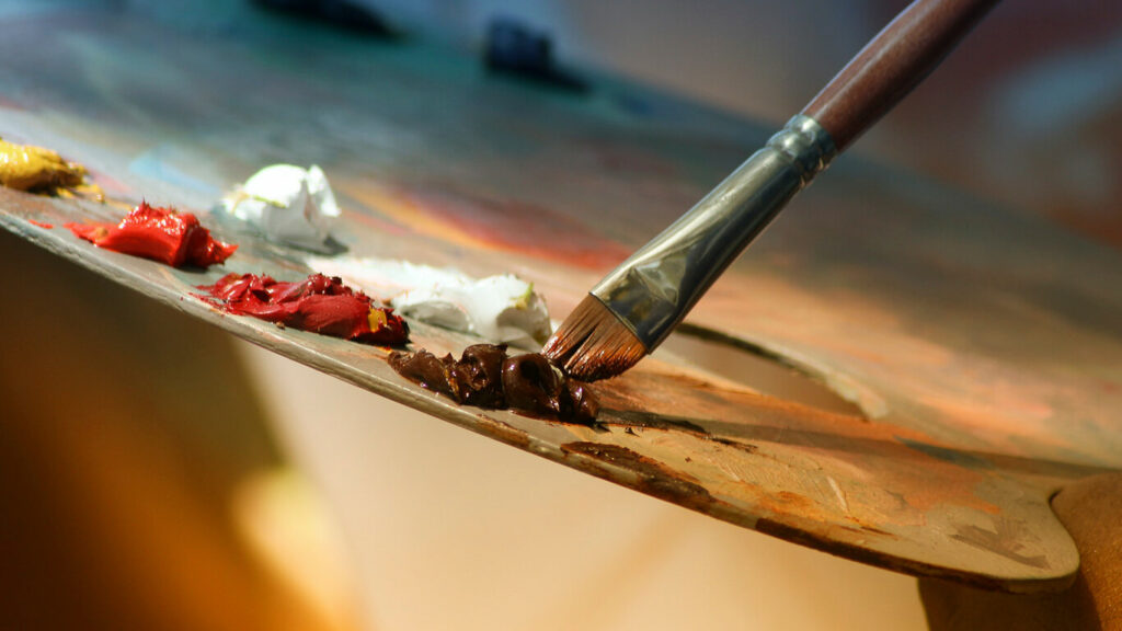 Uma mão de alguém experimentando um dos hobbies para desenvolver - pintura e desenho - pincelando uma paleta de cores.