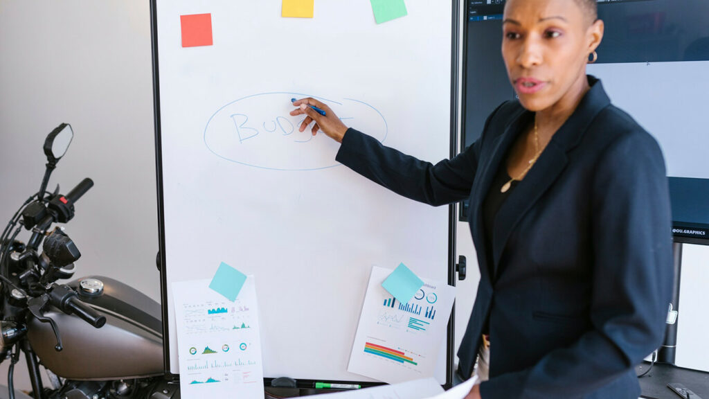 Mulher apontando para uma lousa que tem blocos adesivos colados e aplicando o método Kanban.