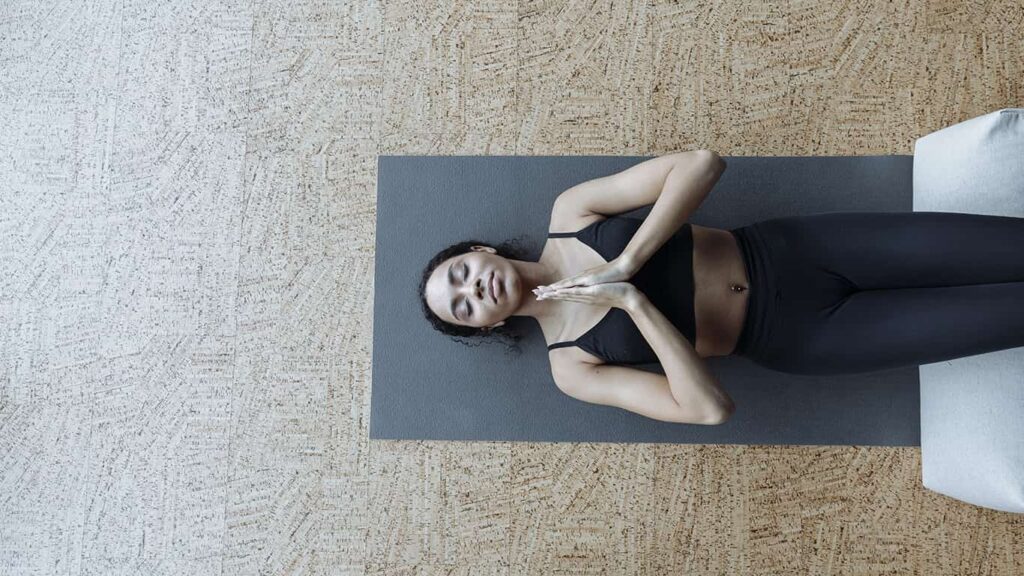 Mulher deitada em um mat, vista de cima, com as mãos juntas e os olhos fechados, em uma meditação, dica da palestra sobre saúde mental de Mari Luz.