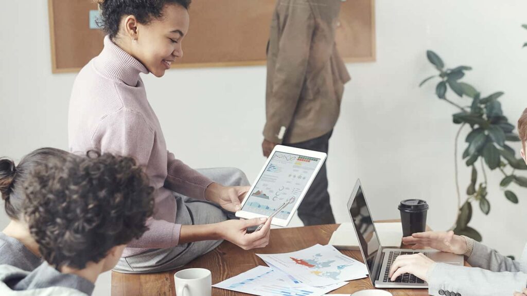 Mulher sentada sobre uma mesa com um tablet na mão, mostrando gráficos e dados produzidos a partir da definição da Matriz RACI para duas pessoas sentadas em cadeiras.