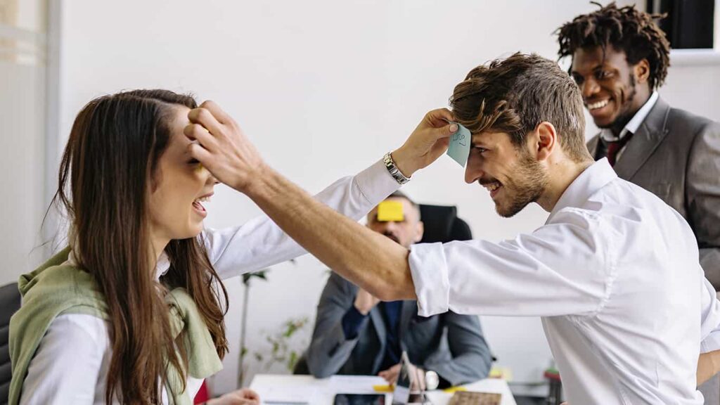 Uma mulher e um homem brancos e de roupas formais de trabalho colocam post-its um na testa do outro, sorrindo, enquanto são observados por outros dois homens também vestidos socialmente para o trabalho em uma experiência de edutretenimento.