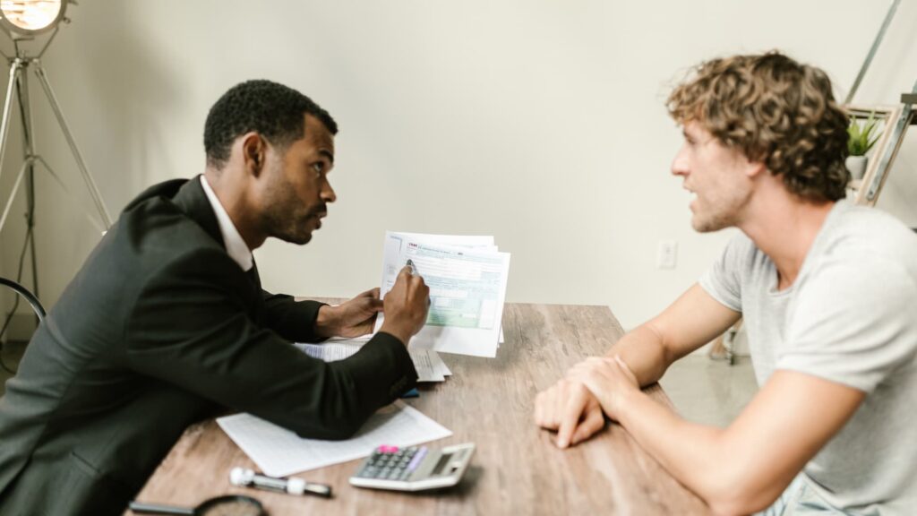 Dois homens sentados ao redor de uma mesa. Um deles segura papéis e caneta e tem uma calculadora a sua frente, discutindo sobre ratings e investimentos.