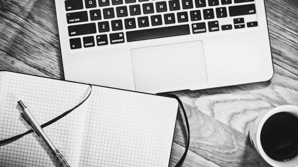 Notebook aberto em cima de uma mesa ao lado de um cadenor de anotações quadriculado.
