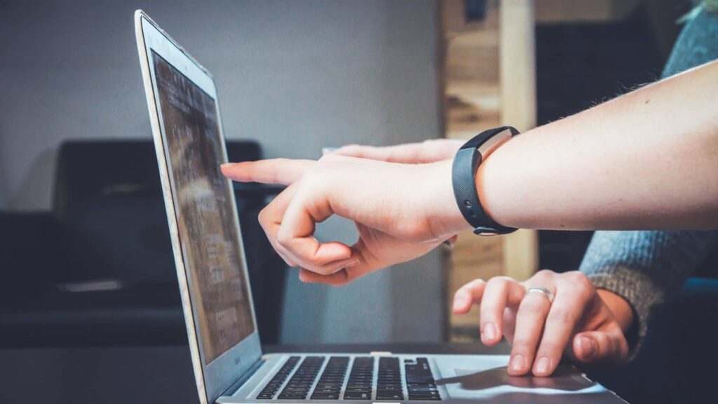 Hand pointing to a laptop's screen while another person's hand uses the touchpad.