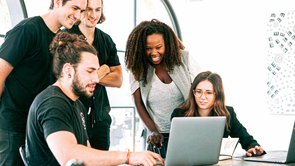 na foto, claramente nota-se o papel do gestor de vendas em uma equipe ética ao ensinar fundamentos aos demais integrantes na mesa enquanto olham um notebook.