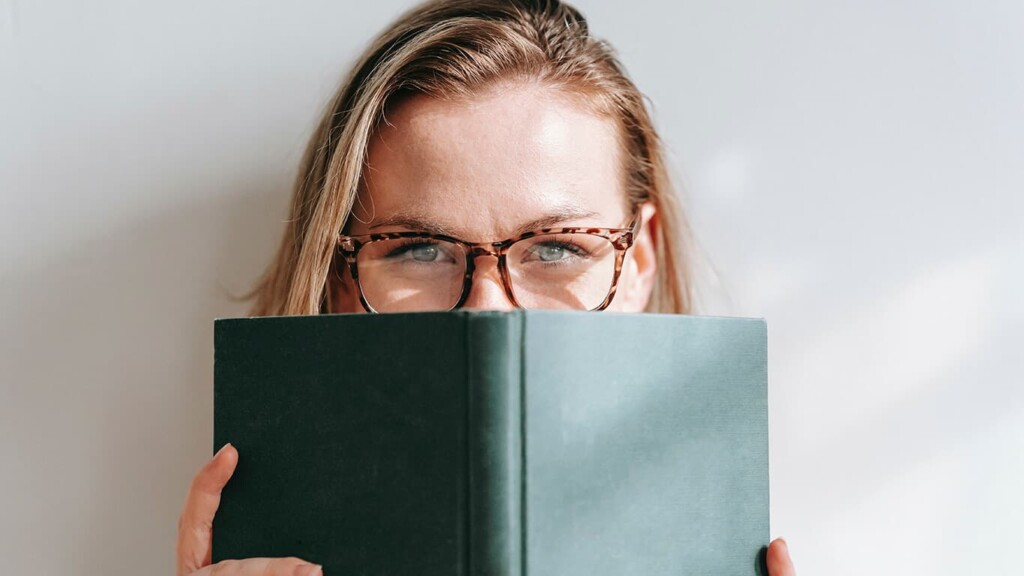 Mulher loira segurando um livro em metade de sua cara representando que, para um uso saudável e consciente do celular, muitas vezes é preciso não usá-lo.