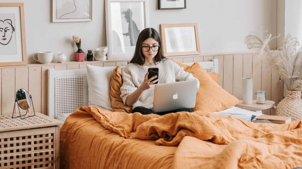 Mulher sentada na cama com um notebook em seu colo e um celular na mão.