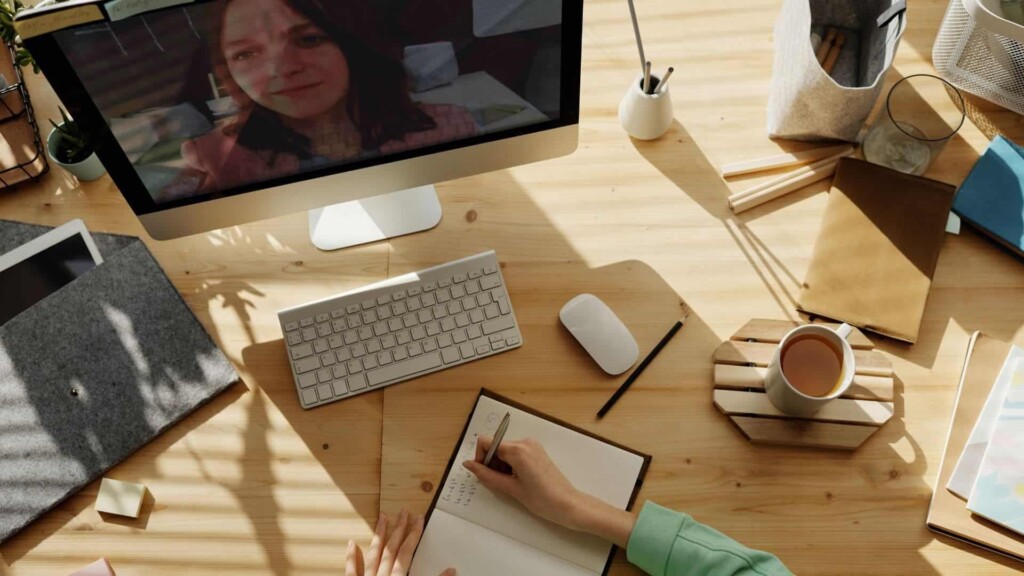 Woman sits at a table while watching someone on her computer screen and writing something on her notebook.