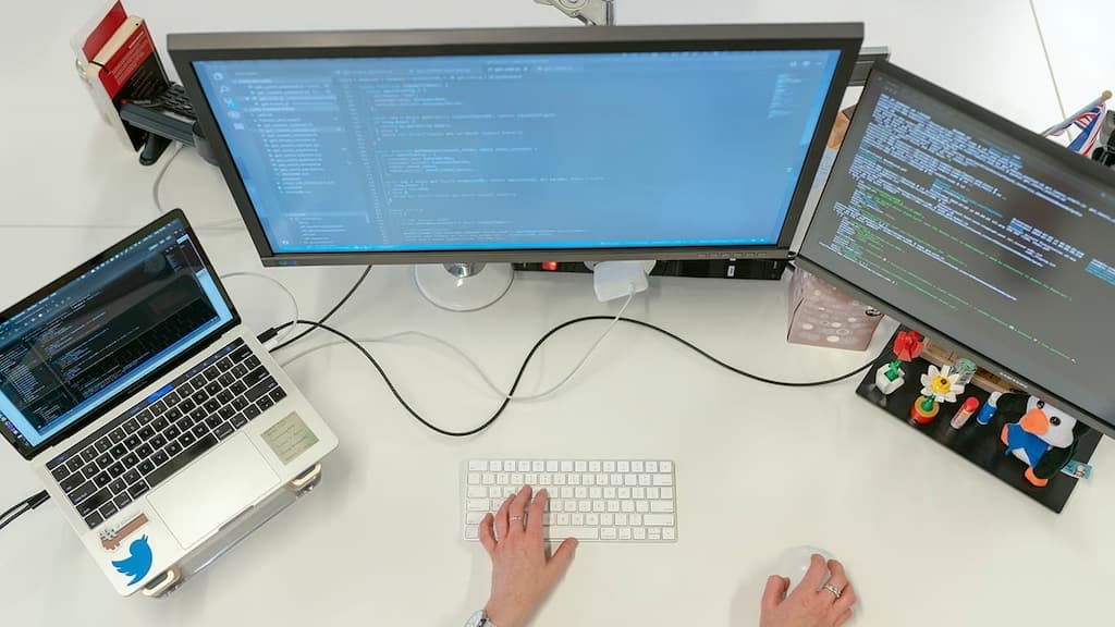 Três computadores vistos de cima, trabalhando com Engenharia de Dados. Uma mão no teclado e uma mão no mouse.