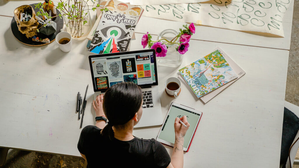 Pessoa em frente a um computador, com diversos objetos ao lado em cima da mesa, para simbolizar o desenvolvimento de habilidades ao cursar MBA em Marketing.
