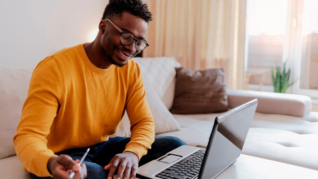 Homem com suéter amarelo sorrindo e olhando para o notebook, como se estivesse desenvolvendo um TCC.