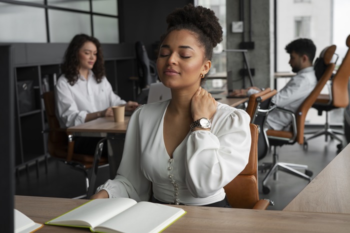 Três pessoas estão em um escritório aconchegante; ao fundo, vê-se uma mulher e um homem, trabalhando tranquilamente; no primeiro plano, uma jovem profissional está sentada à mesa, com caderno e monitor à sua frente, fazendo um alongamento de pescoço.