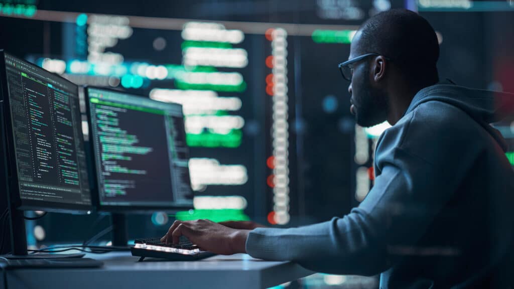 Homem negro, careca, usando um moleton cinza claro, sentado em uma mesa de computador com dois monitores, em um cenário cheio de dados de programação.