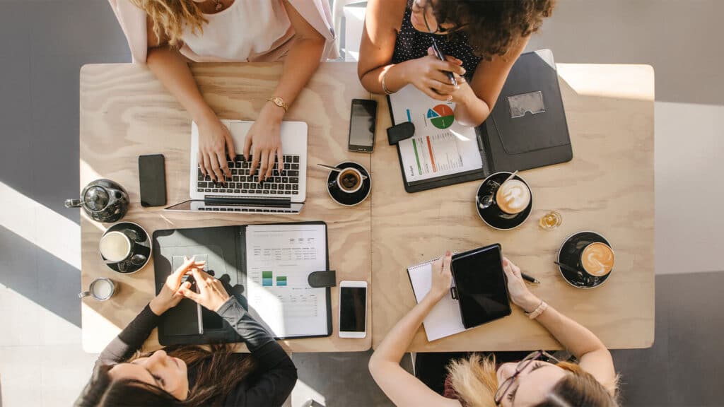 A foto proporciona uma visão de cima da mesa de trabalho, onde quatro pessoas estão sentadas com celulares, computadores, cadernos e outros aparelhos em frente enquanto conversam, para simular uma interação entre as equipes de vendas e marketing.