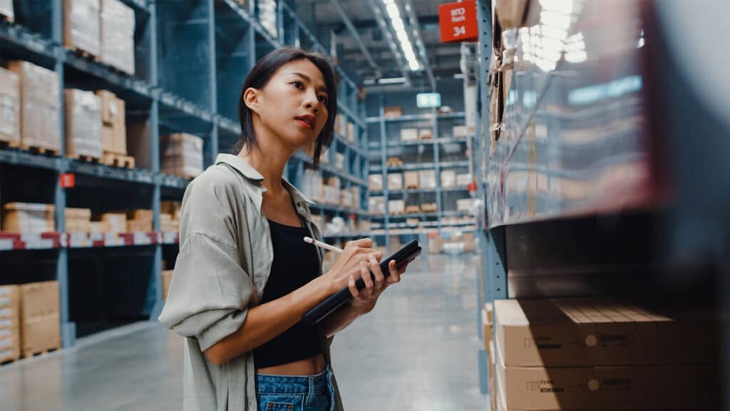 Uma pessoa está entre várias caixas em prateleiras enquanto usa um tablet, em relação às tendências da NRF 2024, o maior evento de varejo do mundo.