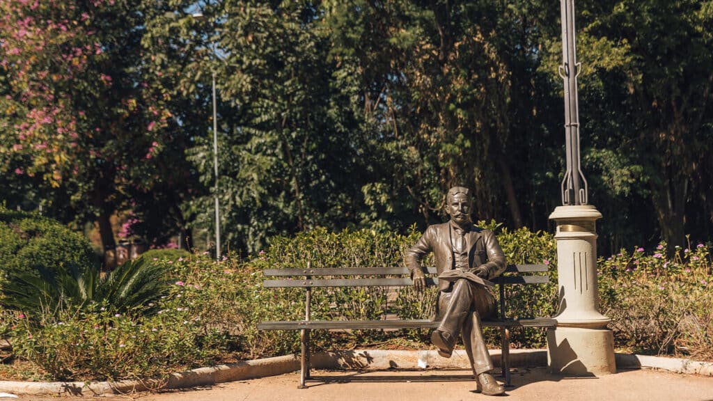 Imagem de uma parte do campus da Escola Superior de Agricultura "Luiz de Queiroz", onde existe um banco com a estátua do Luiz de Queiroz sentado. Os participantes do MBX - Master Business Experience visitam o local. 