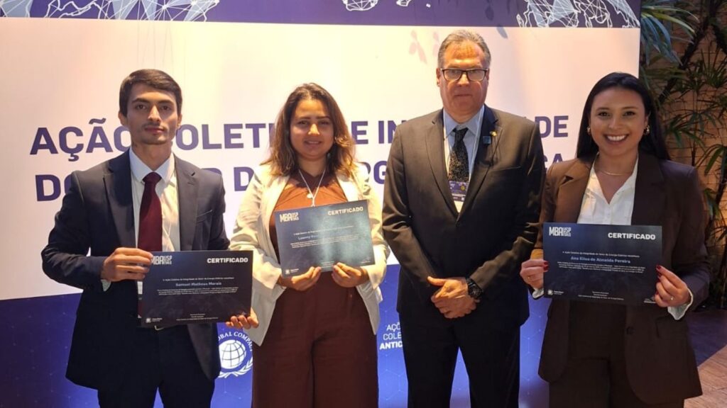 Imagem de alunos do MBA USP/Esalq, premiados no concurso de redação, segurando uma plaquinha do Pacto Global da ONU. Eles estão sorrindo, em um ambiente formal de evento. A imagem transmite o reconhecimento e a celebração pela participação no evento, simbolizando o compromisso com a integridade e compliance no setor de energia.
