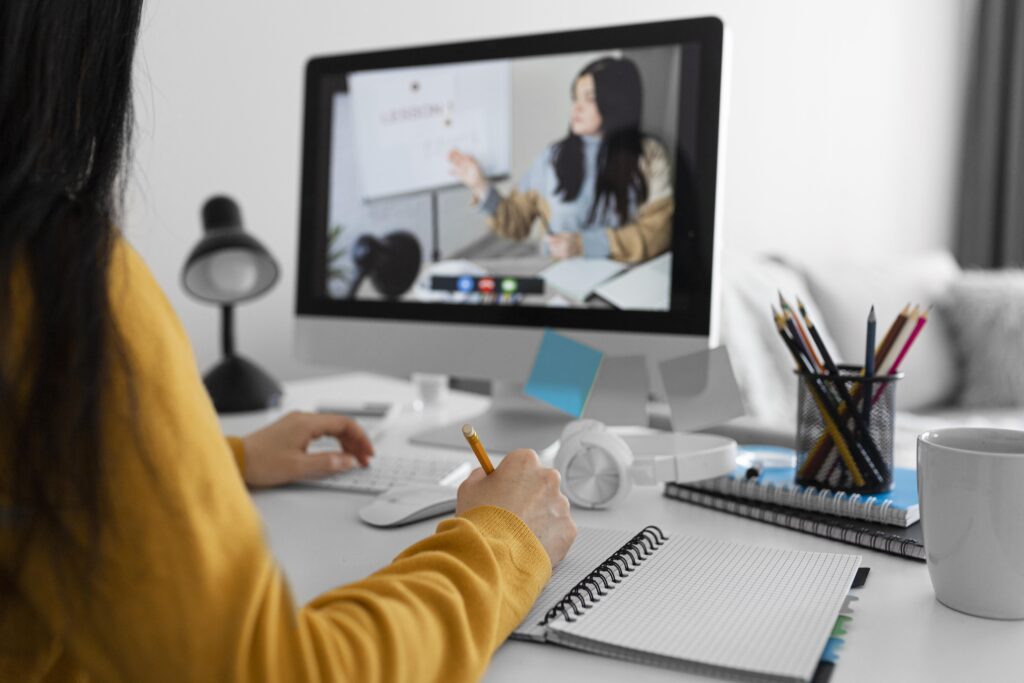  Mulher estudando em frente ao computador; ela assiste aulas online e faz anotações em um caderno.
