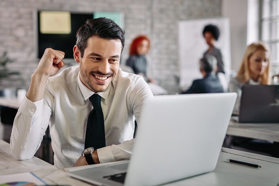 Em um ambiente corporativo, um jovem homem branco comemora uma conquista profissional após aprender como se destacar no trabalho; ele usa camisa social branca e gravata preta, e está em frente a um computador.