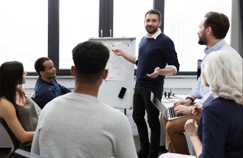 Equipe reunida enquanto um líder apresenta ideias no quadro branco, ilustrando na prática como ser um bom líder ao comunicar com clareza e engajar o time.