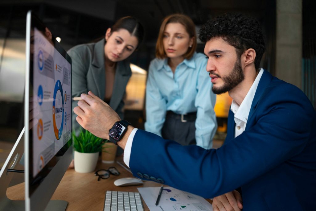 A imagem mostra um profissional explicando gráficos e dados em um computador enquanto dois colegas observam atentamente, destacando um momento de análise estratégica e colaboração em equipe.