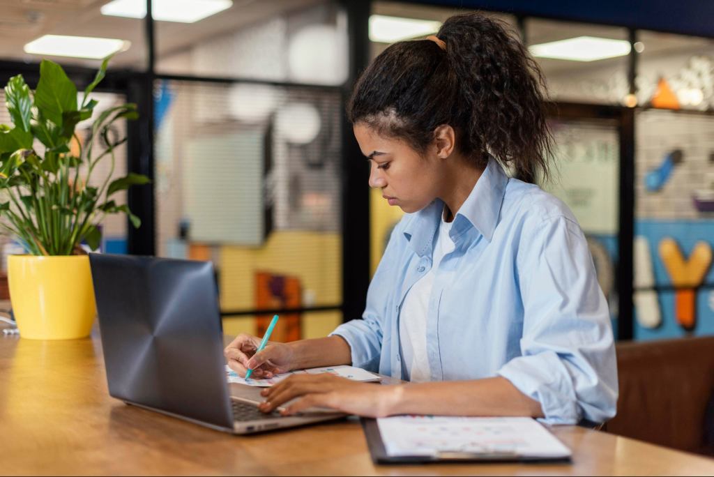 Imagem mostrando uma mulher analisando e escrevendo em uma folha de papel, em um escritório, com o laptop em cima da mesa, sua expressão é pensativa.