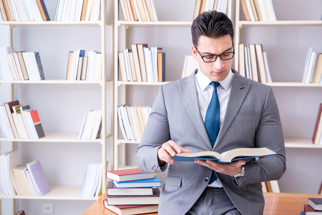 Um homem jovem de terno cinza e gravata lê atentamente um livro em uma biblioteca, com prateleiras repletas de obras ao fundo. A imagem ilustra a dedicação acadêmica e a profundidade de conteúdo oferecidas nas diversas opções de cursos no MBA executivo.