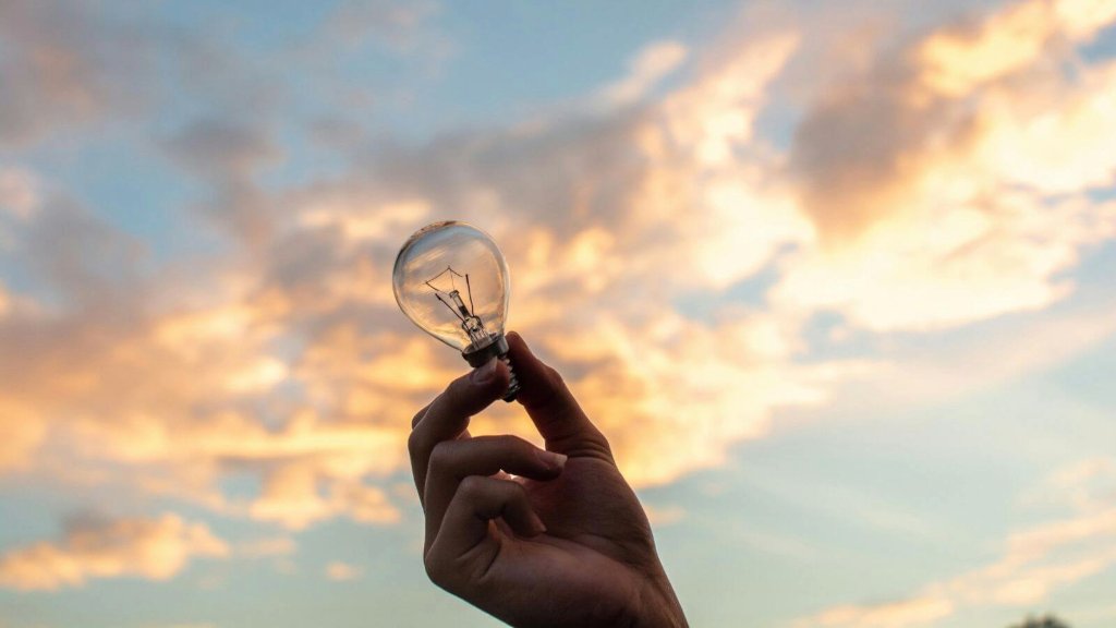 Mão segurando uma lâmpada acesa simbolizando ideias e profissões do futuro diante de um céu iluminado