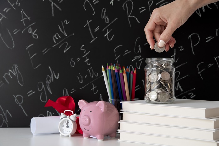 Close-up em objetos relacionados a finanças e educação; há livros e cadernos empilhados, lápis coloridos, um certificado preso com fita vermelha, um relógio analógico, um cofrinho tradicional com formato de porquinho e um pote de vidro onde moedas estão sendo colocadas; ao fundo, há uma lousa com equações matemáticas.